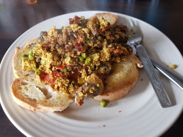a plate with scrambled tofu on toast, the tofu is yellow and there are pieces of tomato, mushroom, and peas