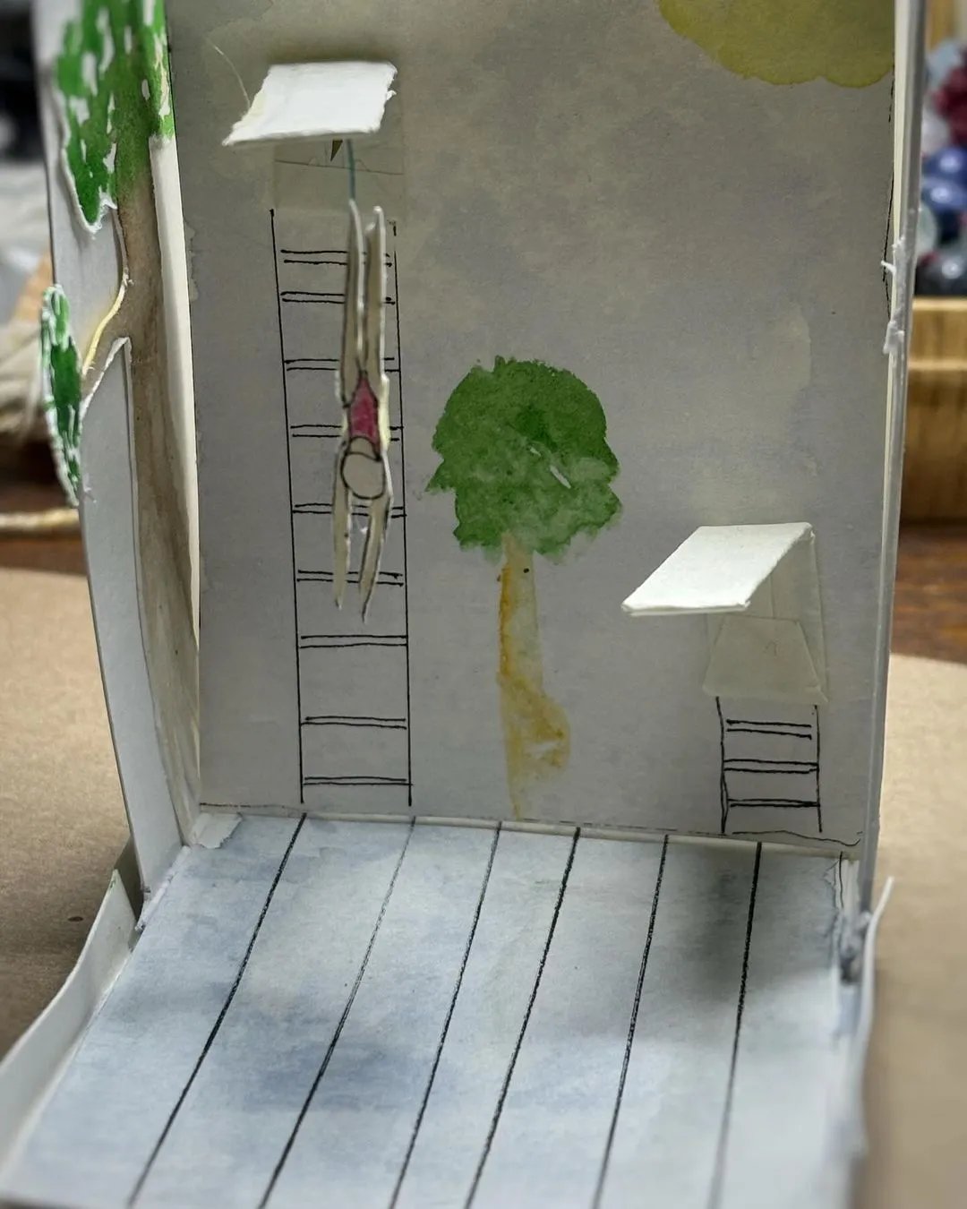 Image of a small diorama of a pool with two diving boards and a figure in a red bathing suit diving from the high board. Image of a small diorama of a pool with two diving boards and a figure in a red bathing suit diving from the high board.