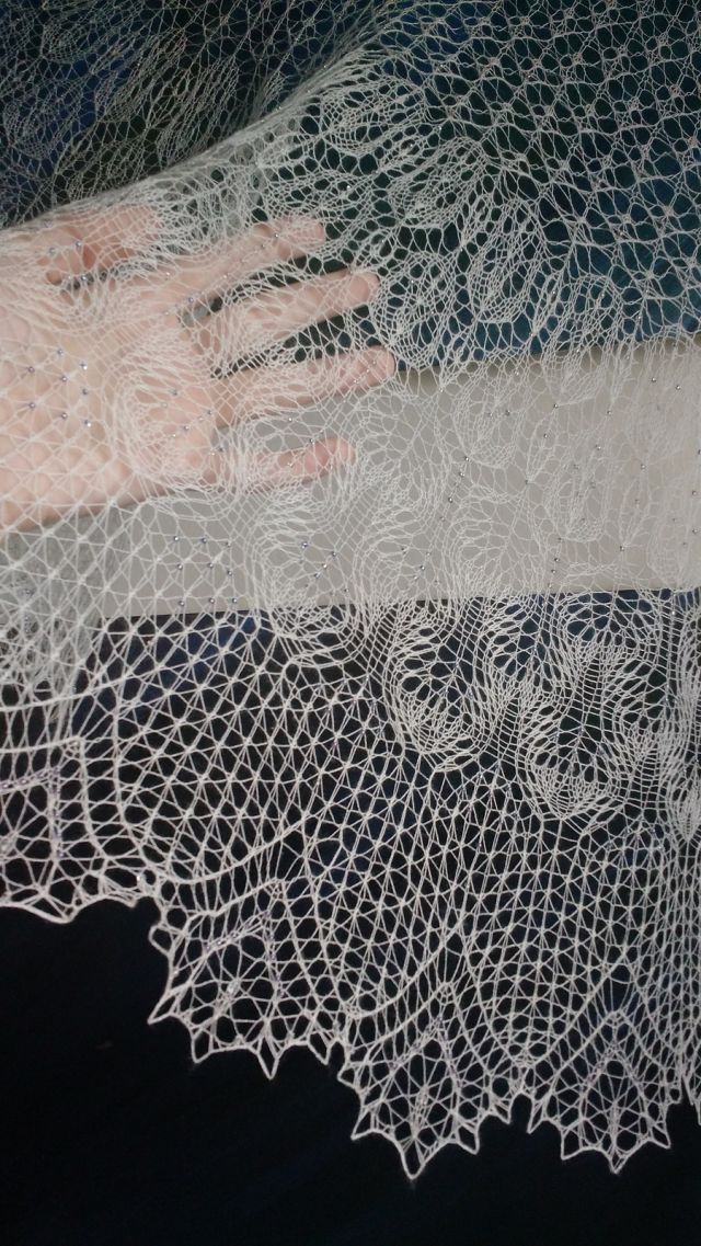 knitted lace so fine it's mostly transparent, sparkling with beads, my hand and the wall showing through behind it. the pattern is undulating and organic, shifting to a more geometric edge.