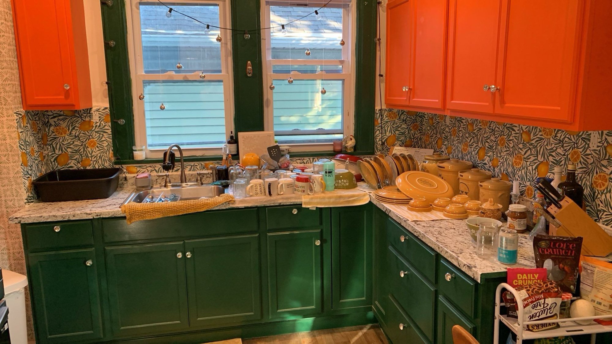 Jozef and Emory’s kitchen, with clean dishes drying on the counter. The upper cabinets are bright orange, and the lower cabinets are a dark green. The backsplash has been covered with a peel-and-stick wallpaper of lemons and leaves (the lemons are sort of orange, though). Disco balls hang across the windows above the sink—we like to keep it fun.