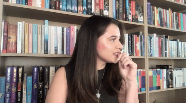 Still from a recording of a video customer interview. A white woman with dark brown hair, wearing a black blouse and a silver necklace, stares off to the right-hand side of the screen, with her hand against her mouth. She looks pensive.