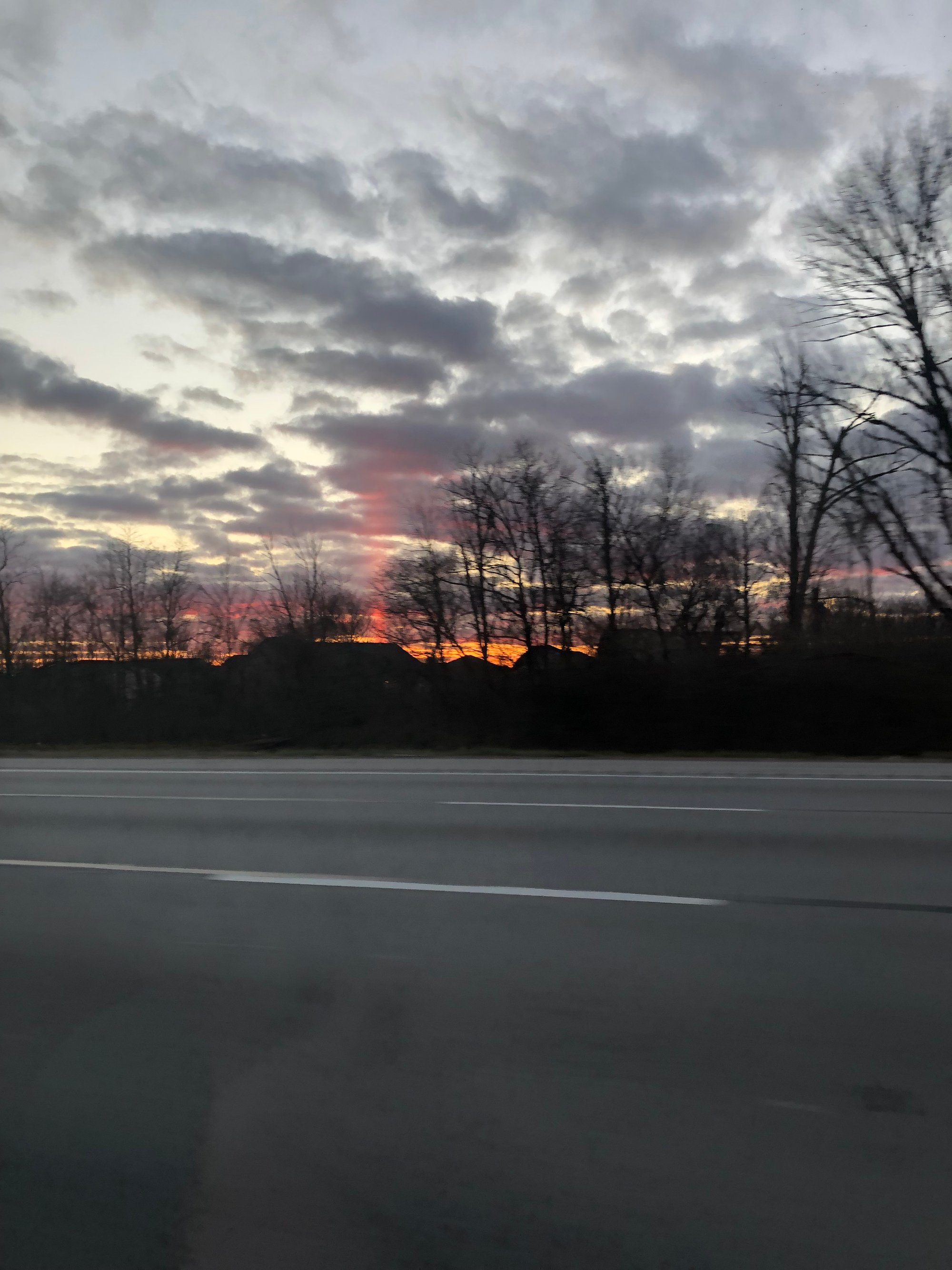 A reddish, magenta-hued beam of sunlight shines down from the clouds in a narrow, nearly vertical line; trees with spidery branches are in front of it, and the highway is foregrounded in front of the trees