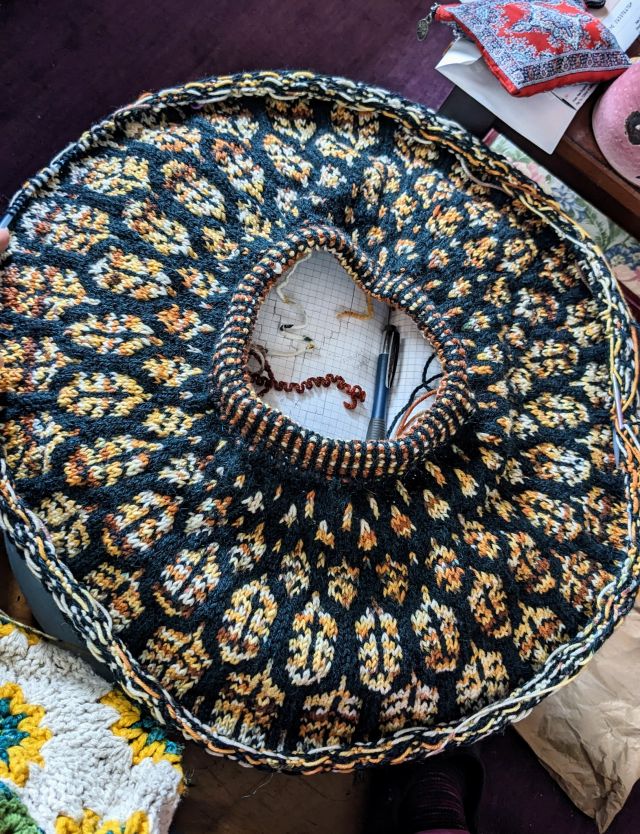 a circle of knitting in progress, a round stripey collar opening in the center, with a pattern of feathers radiating outward from small to large