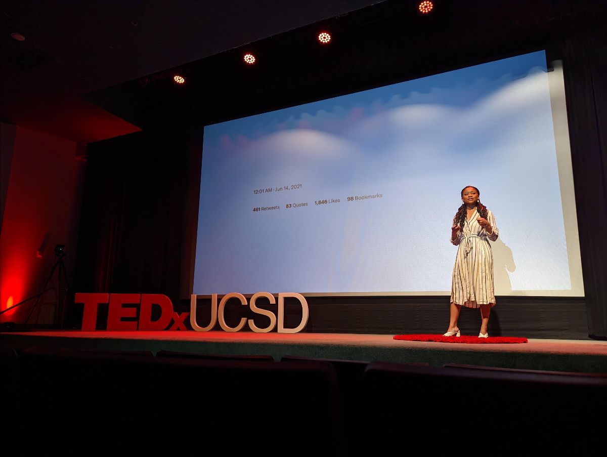 Photo of me on stage in a brown dress. My slides are behind me against the whole backdrop and to the left of the photo, on stage are letters spelling TEDxUCSD.