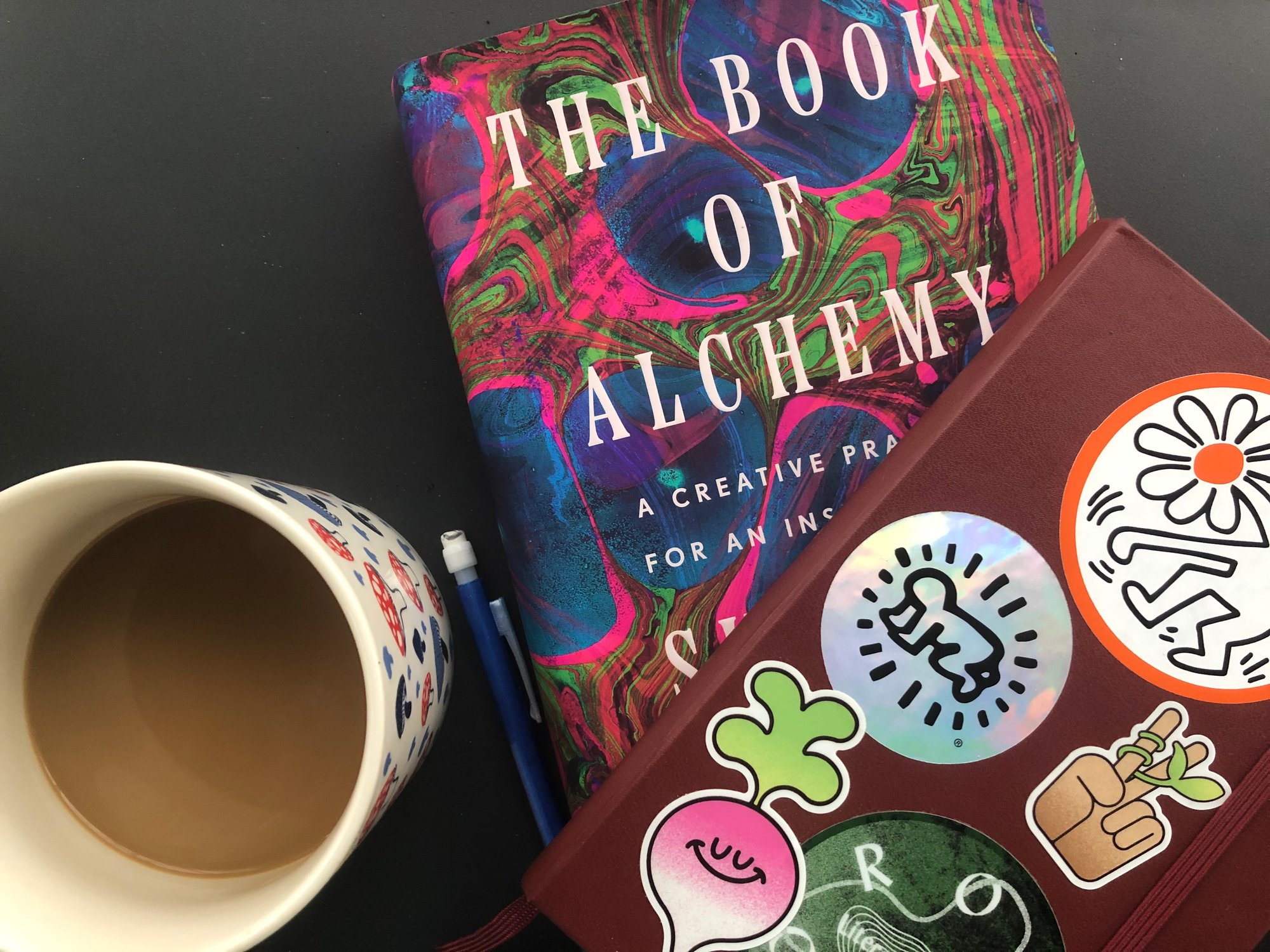 A mushroom-patterned mug half-full of coffee sits on a table next to a copy of Suleika Jaouad’s The Book of Alchemy and Emory’s sticker-covered notebook.