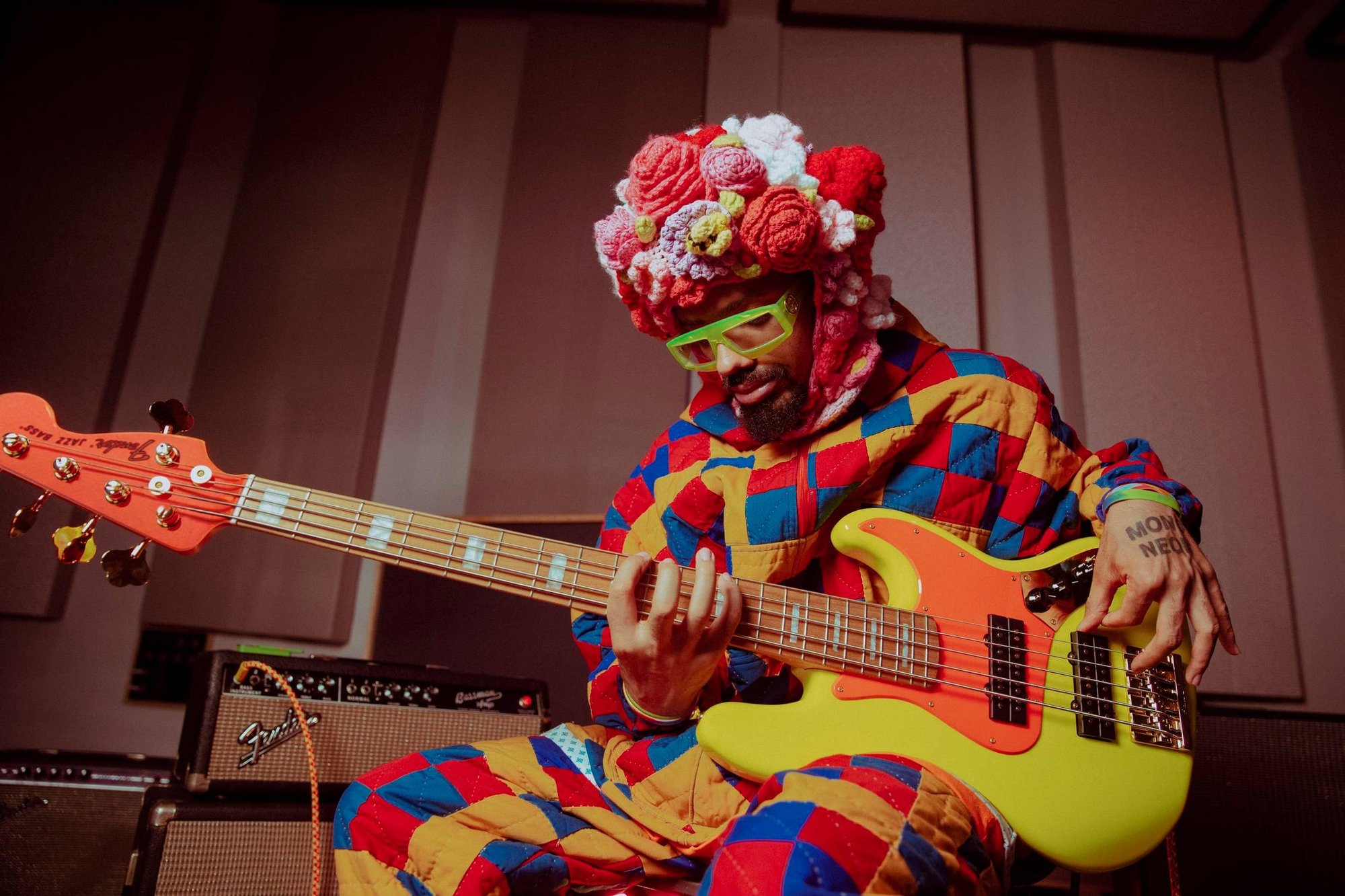 Photo of the artist MonoNeon holding his orange and yellow Fender jazz bass. He is wearing a headdress of knitted flowers, chartreuse sunglasses, and a full-body costume made from a patchwork quilt.