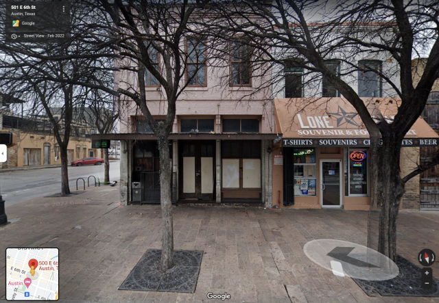 An old storefront building with a metal suspension awning. The windows are boarded up and there's a plastic trash can in front. The building to the right has a fabric awning with the words Lone Star Souvenir and a neon Open sign.