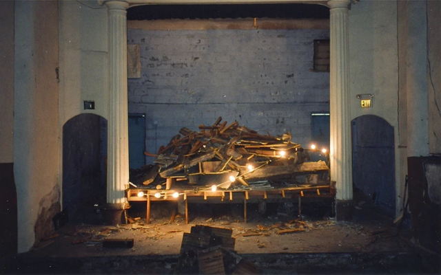 An old, empty stage flanked by two white columns is piled with pieces of wood and debris. A string of bare lightbulbs and one of two exists signs are the only light.