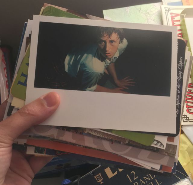 The postcard on the top of the stack is a letterbox photograph of a woman with short wet hair in a white blouse and a plaid skirt, pushing herself up into a sitting position and looking past the left arm of the photographer.