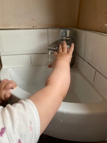 a baby's hand reach for a tap that is on in a small corner sink. The baby is wearing a short white sleeve top with purple flowers on it. He is holding on to the sink with the other hand. The sink has white tiles around it and the walls are bare plaster. 