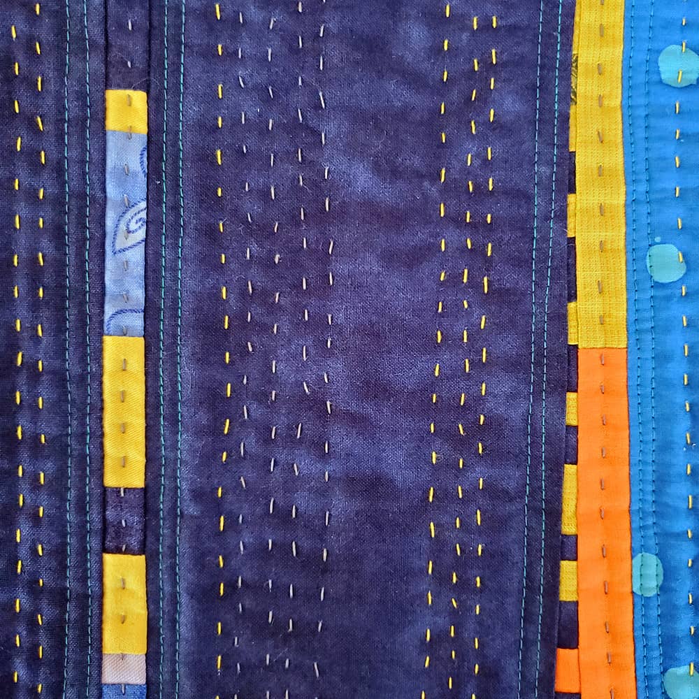 Detail photo of a small patchwork quilt, made with fabrics in blue, yellow, orange, and gray. Both machine-made stitches and hand stitches are visible.