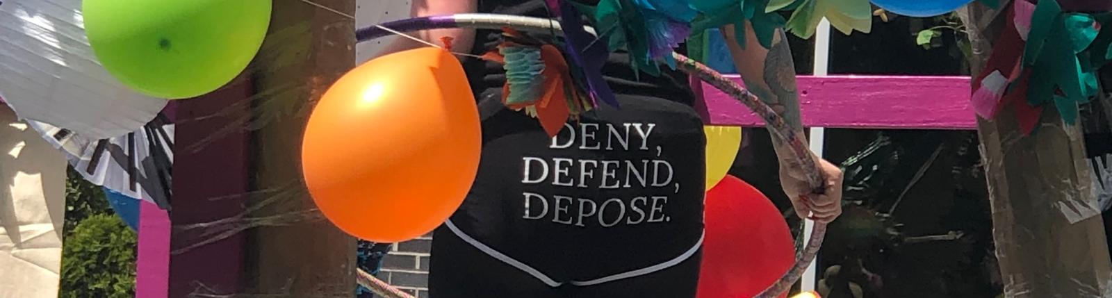 A person holding a hula hoop and wearing booty shorts printed with “DENY, DEFEND, DEPOSE.” The float they’re standing on is decorated with balloons.