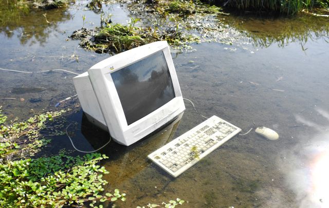 An old desktop monitor and keyboard are posed in a shallow dirty, muddy pond