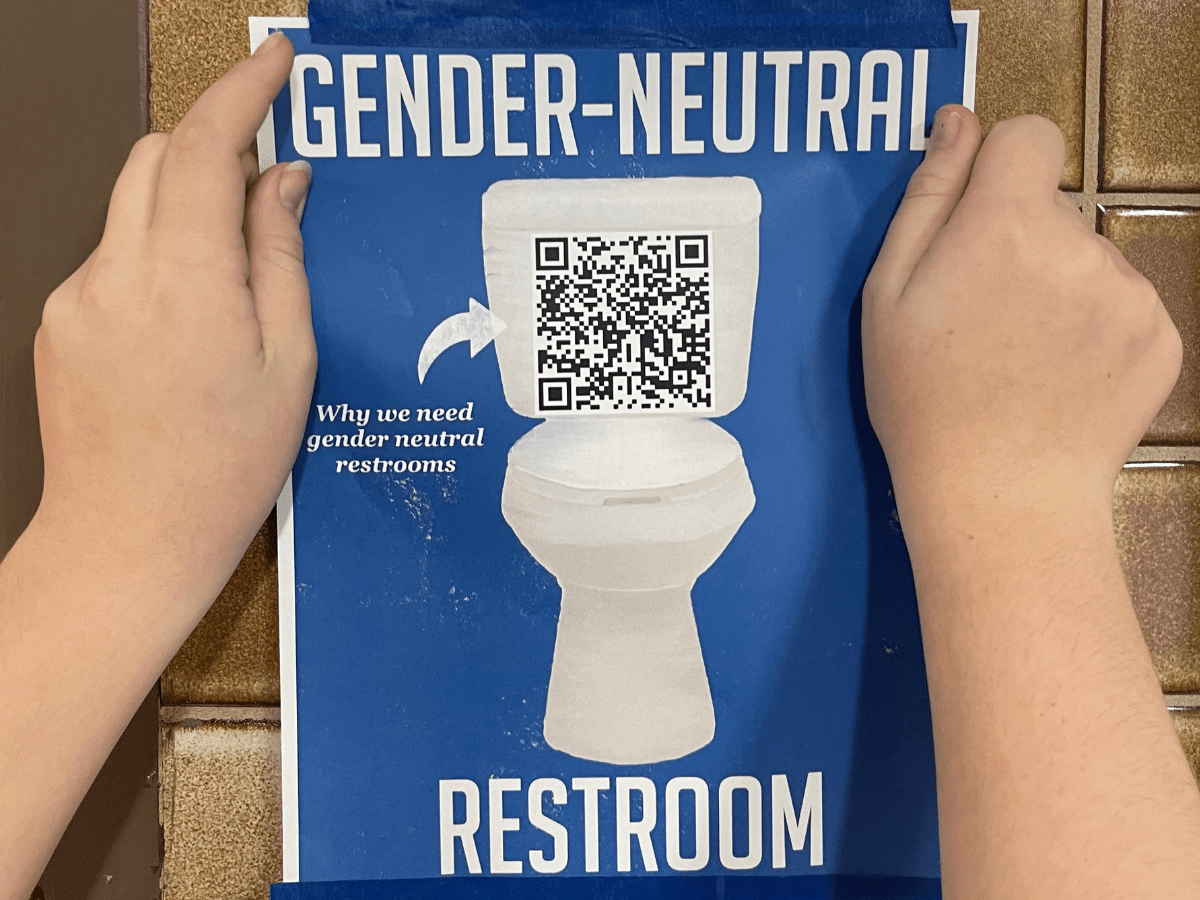 A close-up of hands hanging up a blue sign with a toilet and the words "gender-neutral bathroom" printed on it.