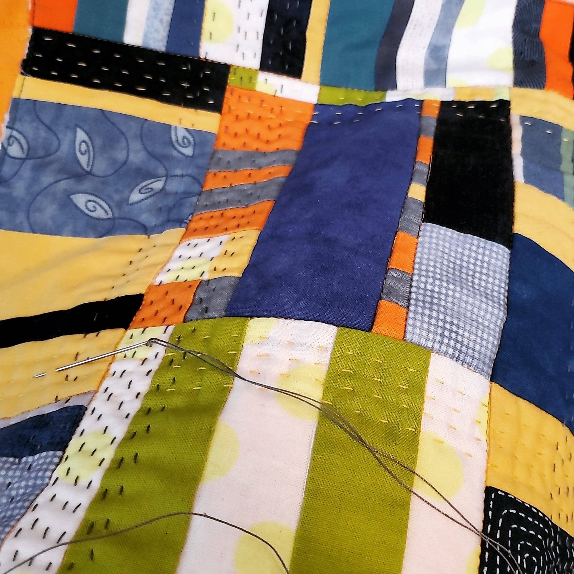 Close-up photo of a small patchwork quilt in progress, with fabrics in orange, yellow, green, dark blue, and other colors. A needle and thread are visible.