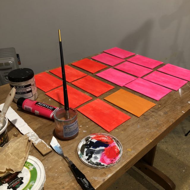 a wooden table holds fifteen small florescent colored canvases waiting to be painted