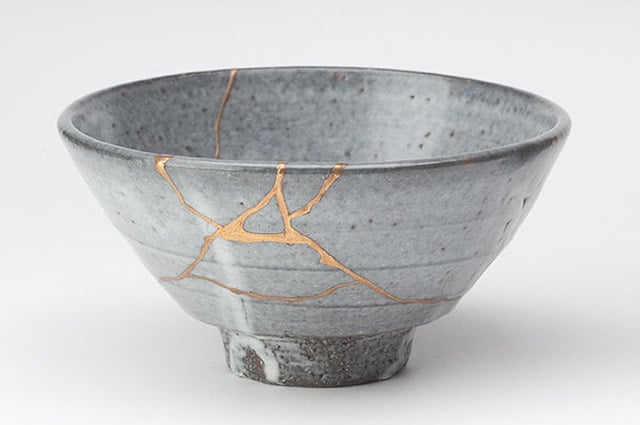 Photograph of a broken grey bowl, reconstructed with cracks filled with gold and resin, forming a noticeable pattern of streams