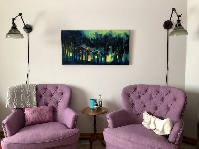 two purple reading chairs with a small round wooden table between are set under a painting of a mystical teal forest scene and two lights. Painting is Healing Forest No. 3 by Carmen Bruhn