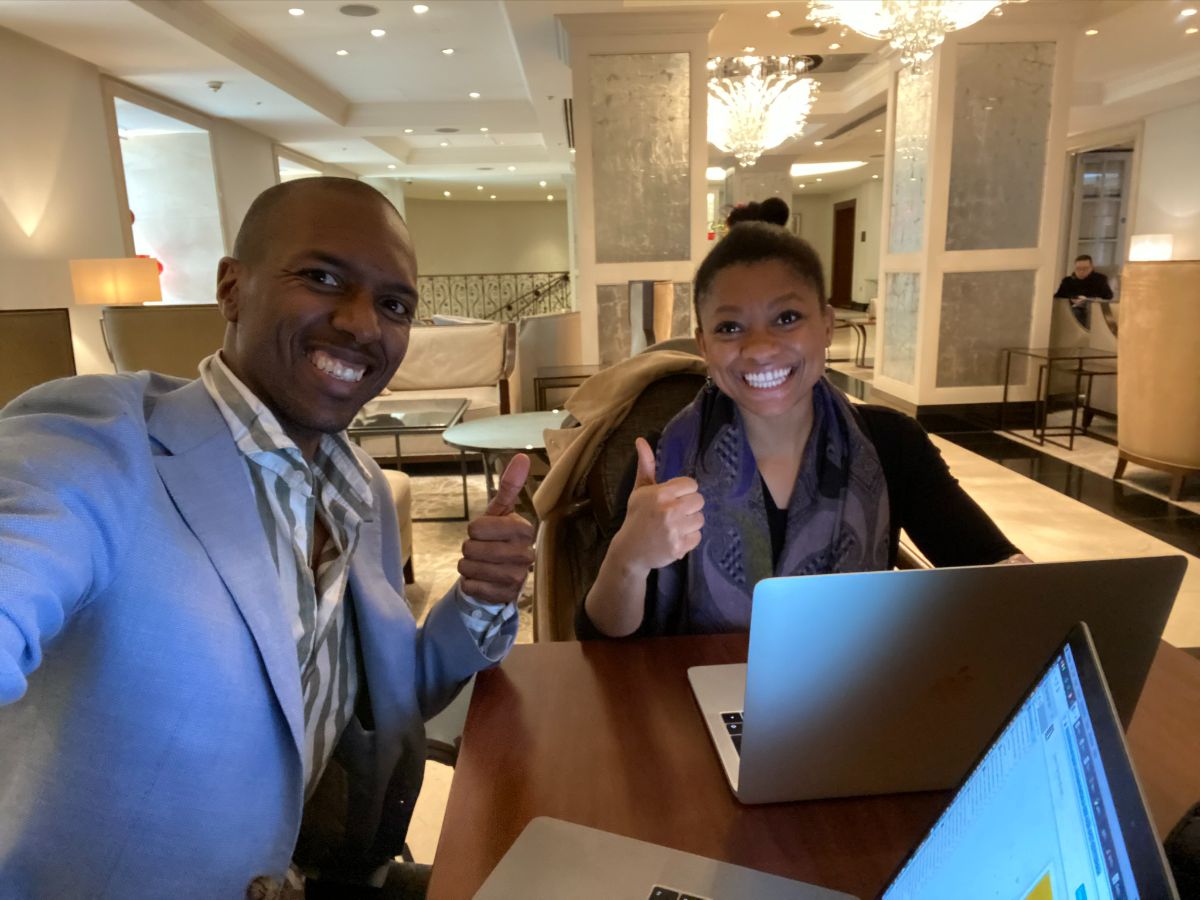 Photo of me and Rob in a hotel lobby area. Rob's arm is extended and then hidden, indicating he is holding up the camera. We are both grinning and holding our thumbs up. On the table in front of us are two laptops.