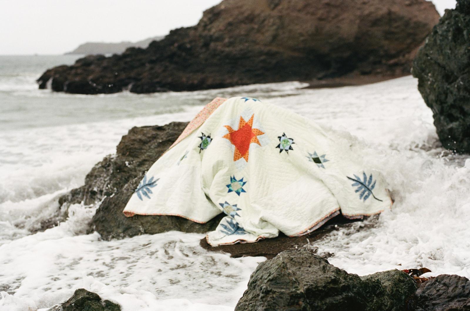 A quilt in shades of green and orange is draped on rocks cropping up from a seafoam-covered beach.