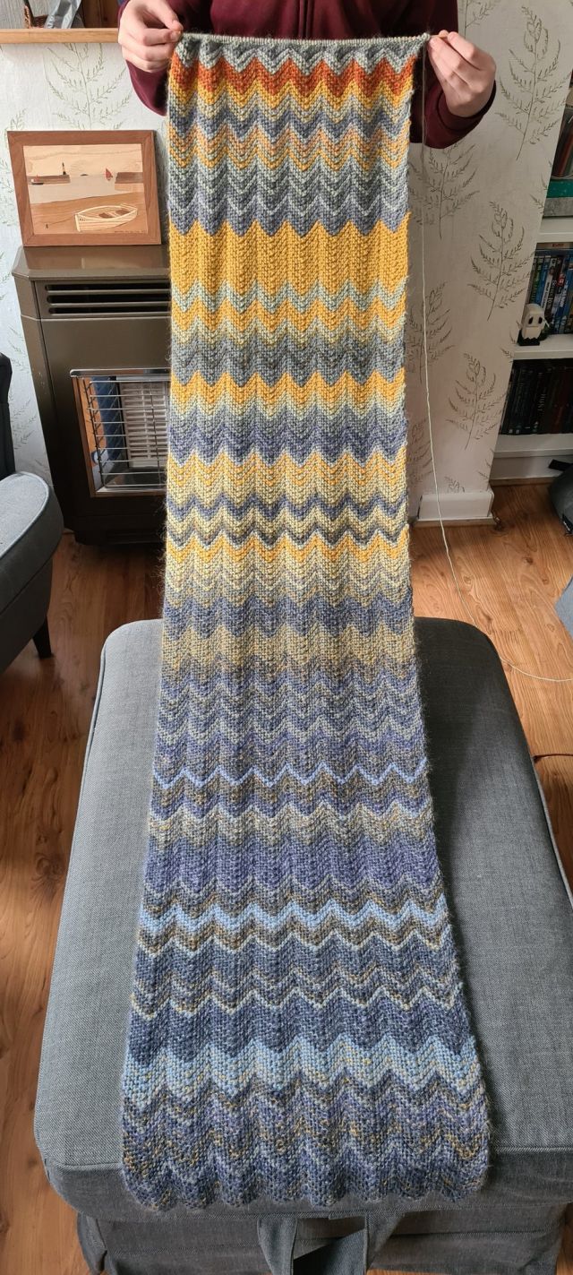 A long hand-knit scarf with rows of alternating colours and materials, going from grey and blue for the winter days to more recent yellows and reds