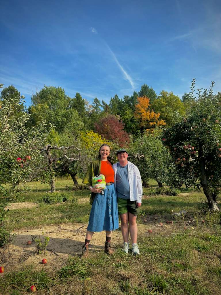Jozef and Emory in an apple orchard, the blue sky bright behind the treeline