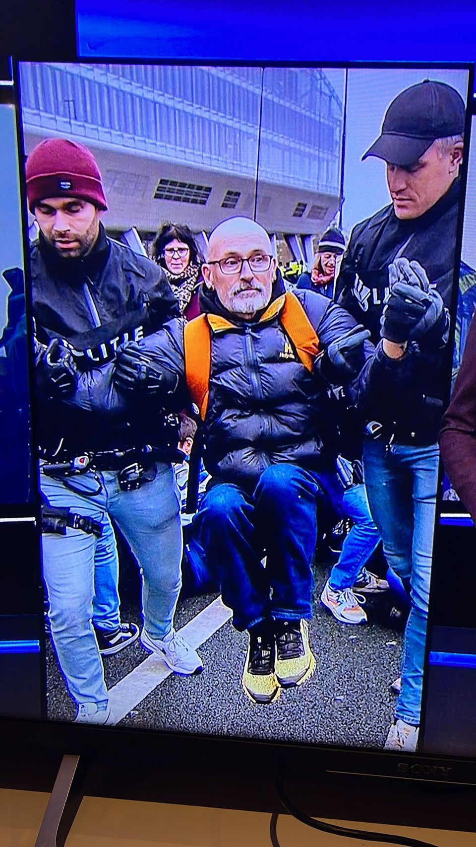 A man with glasses and beard being carried by two poice men.