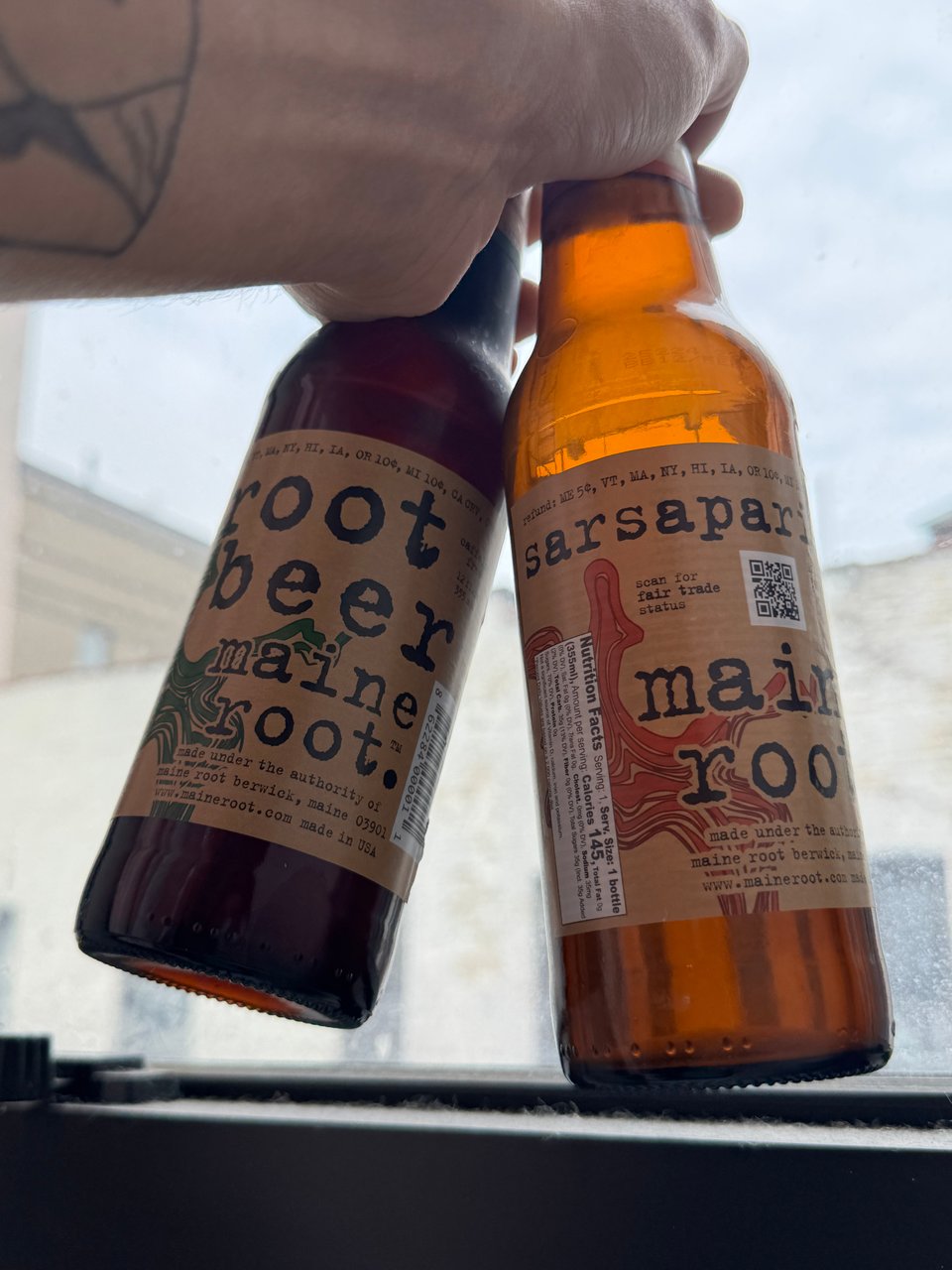My hand holding two bottles, one is Maine Root Root Beer and the other is Maine Root Sarsaparilla.