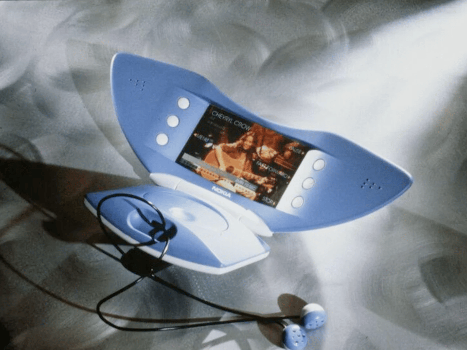 A picture of a very strange-looking phone. It is a clamshell-style flip phone shaped like a large blue crescent. It is opened, showing a colored LCD screen in the middle of the cresent with an image of sheryl crow strumming a guitar. There are two blue earbuds plugged ino the device. There are three white buttons on either side of the screen, and tiny holes in the far left and right of the cresent, indicating that this is where one would speak into the phone and hear the other person talking
