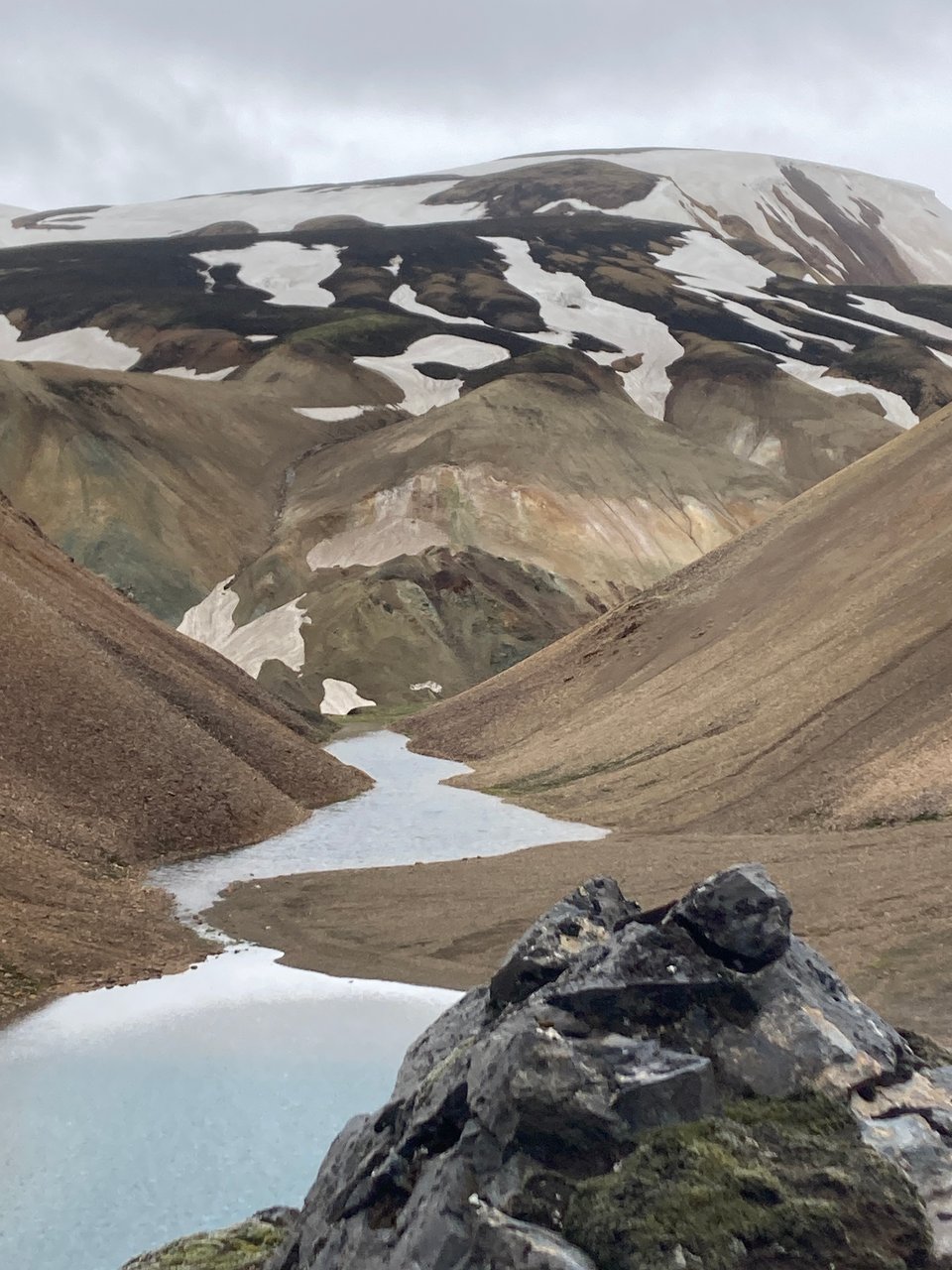 A small river flowing through a valley, against a mountain with some greens and reds, and dotted at the top with snow.