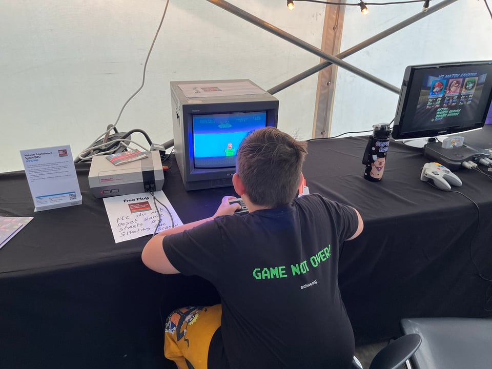 A young boy plays original Mario wearing a black Internet Archive shirt with "Game not over" written on the back.