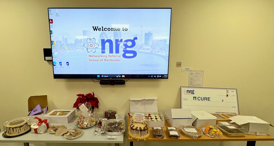 Here’s a shorter version:  **Alt text:** “Tables filled with assorted baked goods for a dessert auction in front of a screen displaying the NRG welcome slide, with a large check for CURE on the right.”