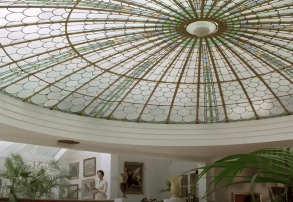 Screenshot from Miss Marple. The main focus is the peacock dome. It's made up of panes of coloured and clear glass shaped like stylised overlapping feathers. Beneath it, the walls have been painted white and have old pictures on them, and there are various palm trees.