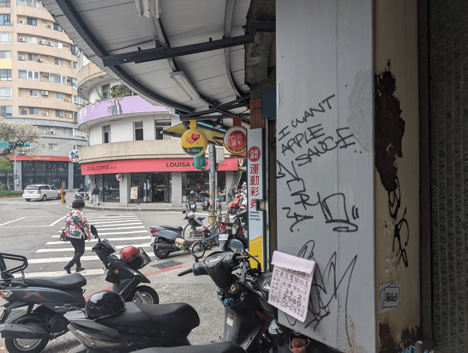 A picture of a sidewalk leading to a crosswalk. The sidewalk has multiple motorcycles parked in it and on the other side of the crosswalk you can see a Louisa Coffee. The focal point of the photo is graffiti on the wall saying "I want apple sauce"