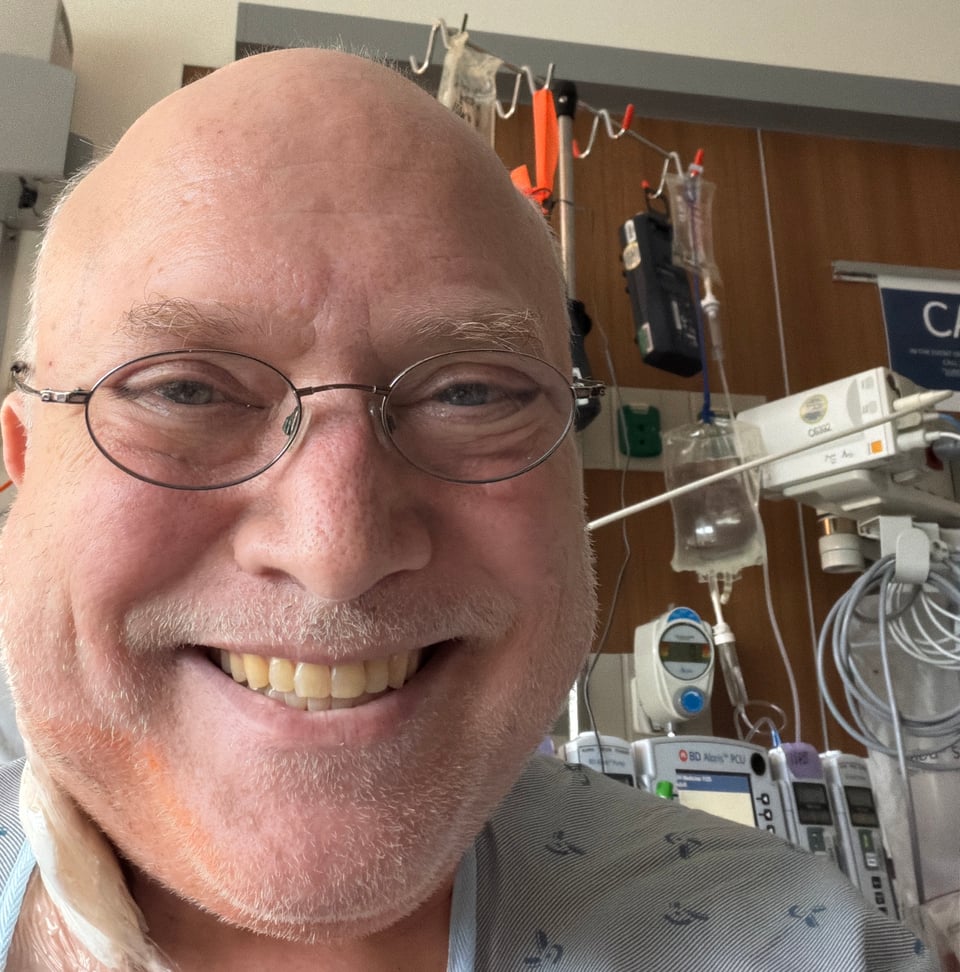 Glenn in a hospital gown from head up, unshaven, wearing glasses, smiling broadly, with various IV bags and other equipment and lines hanging to his left in the background