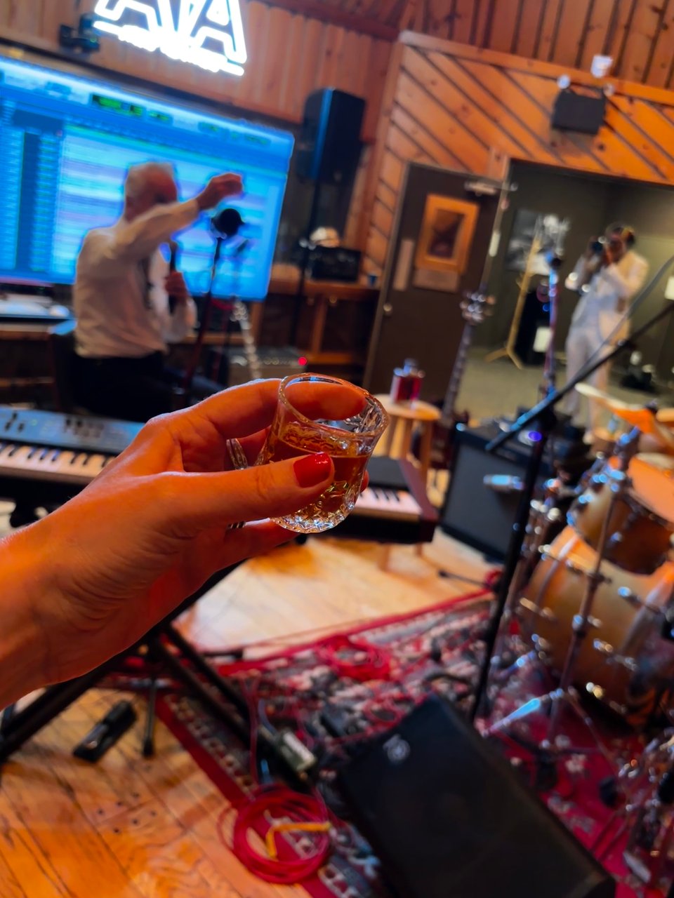 Manicured hand toasting shot of whiskey with guitarist Neil Giraldo in background inside the Power Station Studios, New York City