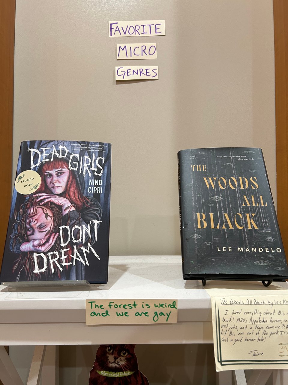 A white bookshelf with a sign saying "Favorite Micro Genres" above it. The shelf shows Dead Girls Don't Dream by Nino Cipri and The Woods All Black by Lee Mandelo. Beneath them is another smaller sign saying "the forest is weird and we are gay."