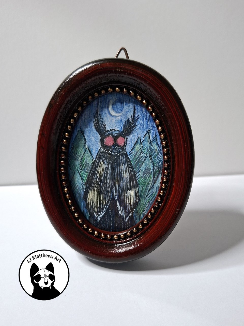 A small oval artwork depicting a whimsical mothman with large ears and red eyes in front of a moonlit mountain landscape, enclosed in a decorative wooden frame.