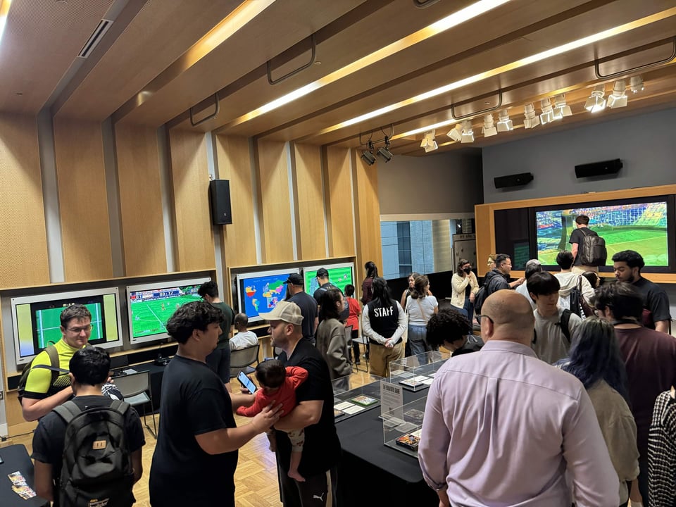 A crowded indoor exhibit space with wood-paneled walls where people gather around several large wall-mounted screens displaying video games, with a glass display case on a black-draped table in the foreground.