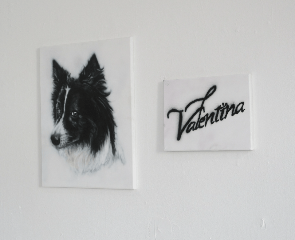 a black and white airbrushed portrait of a collie dog hangs on a white wall beside a painting of the word "valentina"