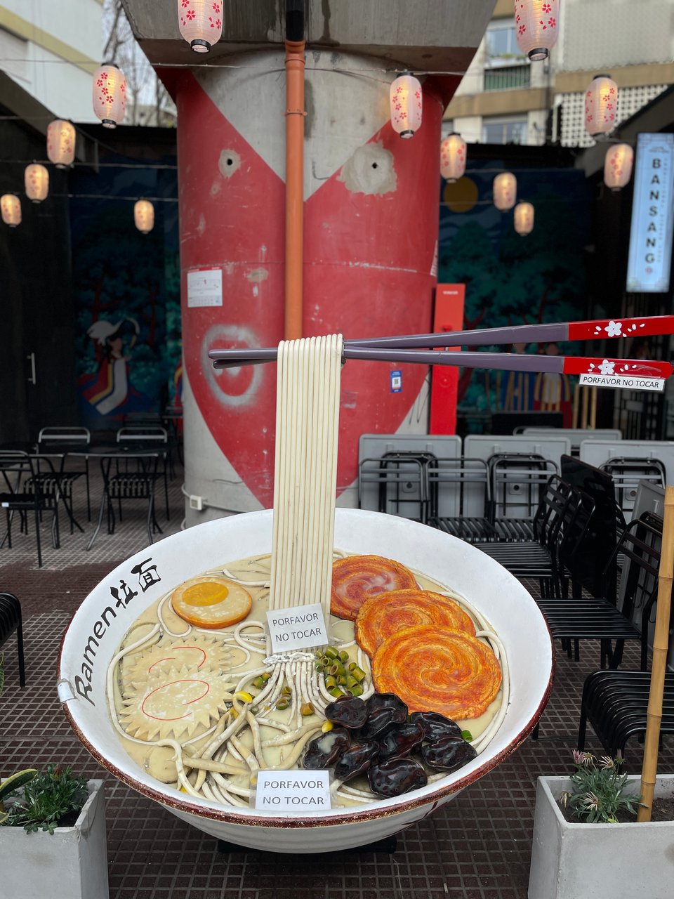 A large sculpture of a ramen noodle soup bowl is on display in an outdoor restaurant.