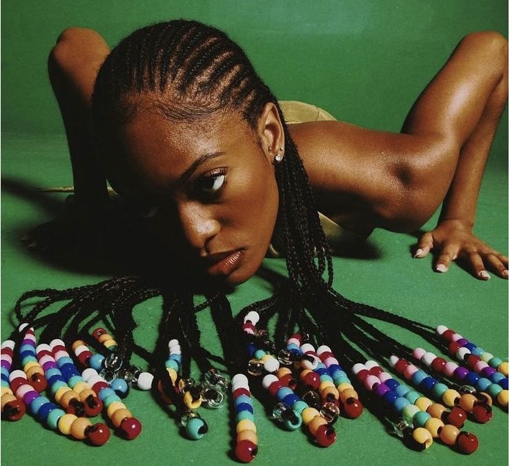 This is a black woman on a green background. Her head is uncomfortably close to the floor and she's looking up at the camera in a way that looks like she knows she's being observed in an uncomfortable situation but also a bit like "so look at me then". She's pushing up against the floor as if she's going to do a push up but her arms are at an angle where it wouldn't be effective. Her cornrows trail onto the floor and have coloured beads at the end of each one.