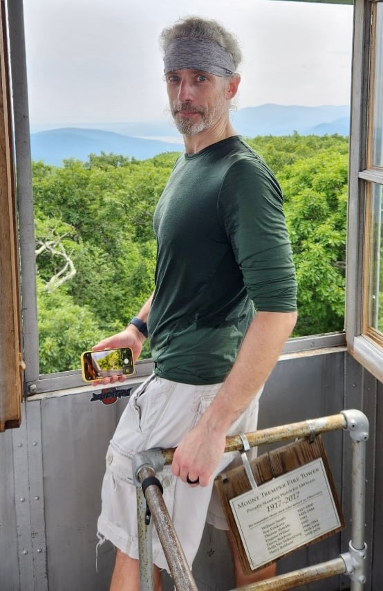 Photo of white man in green shirt, khaki cargo shorts, and gray headband, sweating profusely, atop a fire tower in the Catskills, with trees and mountains behind him