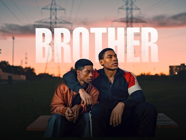 This promotional image for the film Brother features two young Black men seated on a bench at dusk, framed by an orange-pink sunset and silhouetted power lines in the background. One man wears a rust-colored jacket and leans into the other, who wears a black and red tracksuit and wraps his arm protectively around him. The word BROTHER is displayed in bold, white, capitalized letters across the top of the image, partially overlapping with their heads, reinforcing the emotional connection and theme of the film. The mood is intimate and contemplative.
