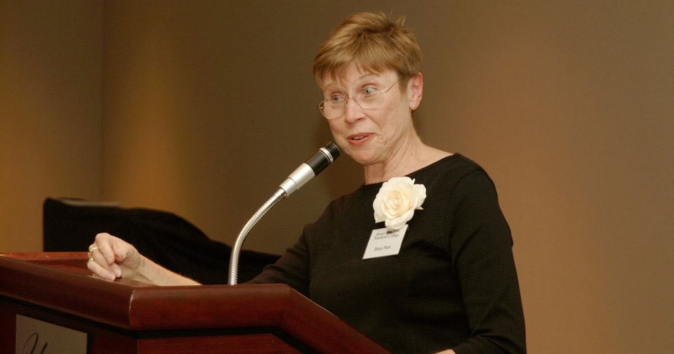 Dixie Platt, with short hair and glasses stands behind a wooden podium, addressing an audience. She wears a black top, has a white flower pinned to her shirt, and speaks into a microphone.