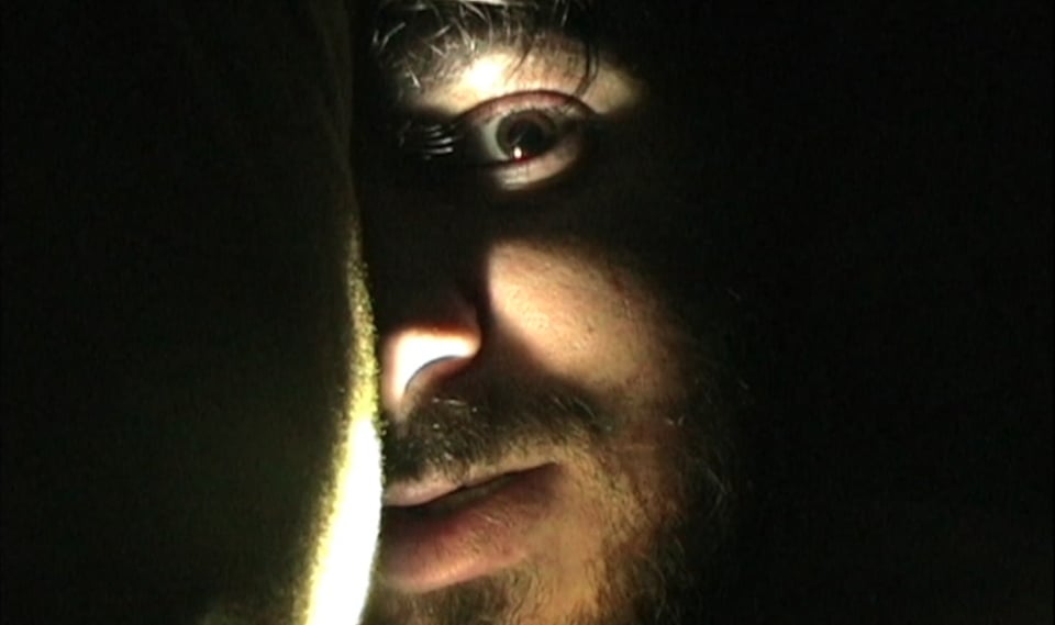 An image from Tinsman Road: a close-up of Robbie in his tent in the woods, his face against a pillow, the light of a lantern revealing only half of his face with one eye staring into the camera.