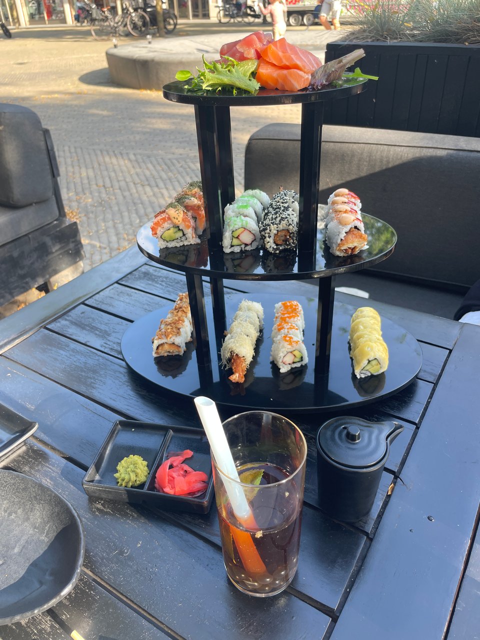 A tower of three plates covered with various sushi rolls. There is a glass of bubble tea in front and a plate with wasabi paste and pickled pink ginger slices.