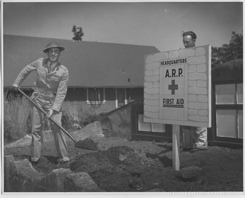 Black and white photo. A man in military uniform with a shovel of dirt. He stands in front of a sign that reads "Headquarters. ARP first aid"