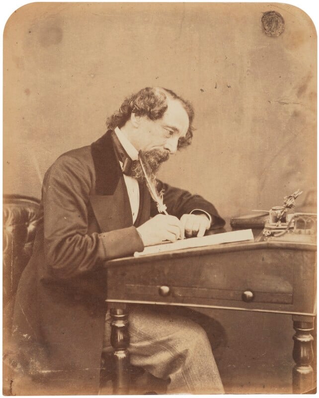 Charles Dickens posing at his writing desk, holding a feather pen and gazing down at his manuscript. He has a large goatee and curled hair.