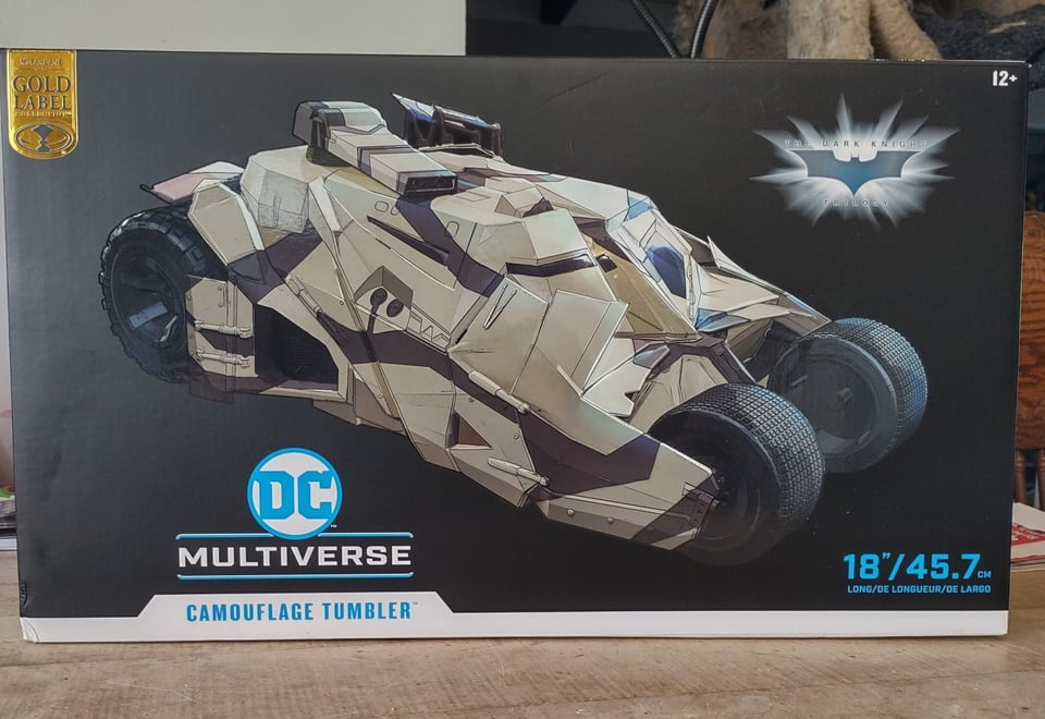 A still-in-the-box Tumbler, the bulky, angular, armored vehicle from the Christopher Nolan Batman movies. This one is the desert camouflage version from the third film. The box is on a wooden table l, lists the size as 18"/45.7 cm long, and has a large DC Multiverse logo on it.